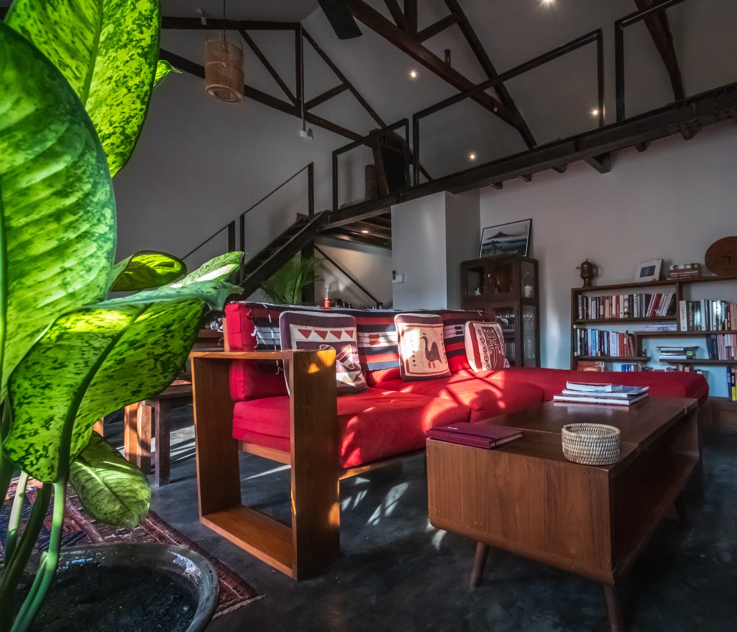 Interior of a restored Sri Lankan heritage penthouse — exposed timber roof trusses, a steel staircase, a red sofa and book-lined shelves
